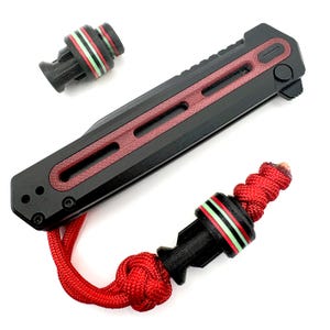 Puede incluir: Cuchillo plegable negro y rojo con un cordón rojo. El cuchillo tiene un mango negro con detalles en rojo. El cordón es rojo y está unido a una cuenta negra con rayas rojas y verdes. También hay una cuenta negra separada con rayas rojas y verdes.