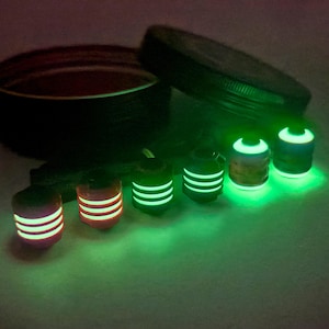 May include: A collection of glowing, cylindrical objects with horizontal stripes, in shades of red, orange, and green. A dark container and lid are in the background. The objects emit a bright, neon green light.