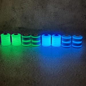 Può includere: Otto rocchetti di filo cilindrici disposti in una fila orizzontale che brillano al buio. I primi quattro rocchetti emettono una luce verde neon brillante, mentre i restanti quattro emettono una luce blu vibrante, mostrando le loro proprietà luminescenti.