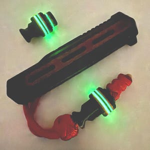 Op de afbeelding: Een zwart en rood multi-tool met gloeiende groene accenten. De tool heeft een getextureerde handgreep en een rood koord. Er zijn ook twee extra stukken met gloeiende groene banden.