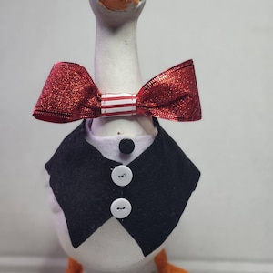 May include: A white goose figurine with an orange beak and feet, wearing a black vest with white buttons and a red glitter bow tie with white and red stripes. The goose has a formal, decorative appearance.