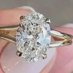 May include: A gold ring with a large, oval-shaped diamond. The diamond is the main feature, with many facets catching the light. The band has a split shank design, giving a contemporary feel to the traditional engagement ring.
