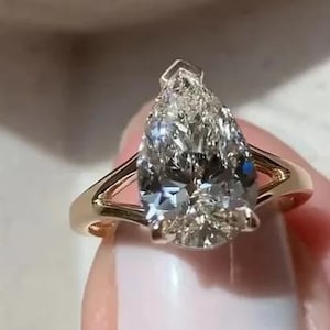 May include: A gold ring featuring a large, pear-shaped diamond. The diamond is clear with multiple facets, and the ring has a simple, elegant design. The band is a warm gold color, and the ring is held up by a hand with white nail polish.