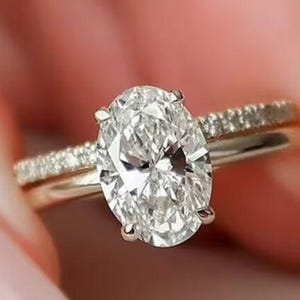 May include: A gold engagement ring featuring a large oval-cut diamond. The band is embellished with smaller diamonds. The ring is held up by a person with a pink manicure.