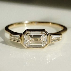 May include: A gold ring with a large, rectangular-cut diamond centre stone, with two smaller, baguette-cut diamonds either side. The ring has a slender band and a modern design, suitable for engagements or special events.
