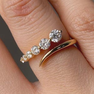 May include: A gold-toned ring featuring a series of graduated, sparkling clear stones. The ring curves around the finger, with the largest stone at the top. The design is elegant and modern, suitable for everyday wear or special occasions.