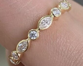 May include: A gold band ring featuring alternating round and marquise-cut clear gemstones. The ring is held between fingers, showcasing the delicate design and sparkle of the gemstones. The ring is a piece of jewelry.