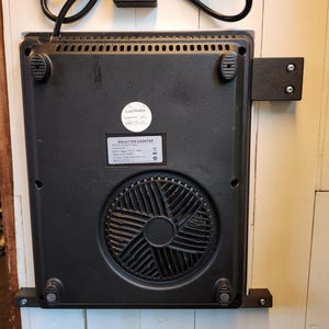 Pode incluir: Placa de indução preta com ventoinha, cabo de alimentação e suportes de montagem. A placa tem um rótulo que diz "INDUCTION COOKTOP".