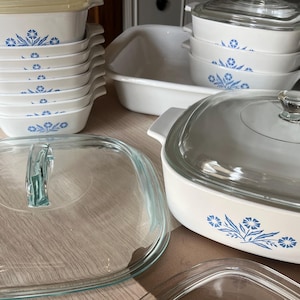 Może przedstawiać: Zestaw białych ceramicznych naczyń do pieczenia z niebieskim wzorem kwiatowym. Komplet zawiera kilka kwadratowych i prostokątnych pojemników, niektóre z przezroczystymi szklanymi pokrywkami, oraz jedno duże prostokątne naczynie bez pokrywki.