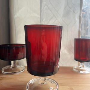 May include: Three ruby red glass goblets with clear stems and bases are arranged on a wooden surface. The goblets have a cylindrical shape and a slightly flared rim. The clear bases provide a contrast to the deep red color of the glass.