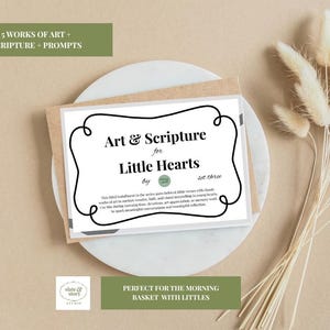 May include: A set of cards titled "Art & Scripture for Little Hearts" with a black border, resting on a white plate. The cards are in a brown envelope. The text includes "5 Works of Art + Scripture + Prompts". Dried wheat stalks are arranged on the right.