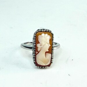 May include: A silver ring featuring a cameo with a profile of a woman. The cameo is set in a rectangular frame with a detailed border. The woman's profile is in a light cream color against a brown background.