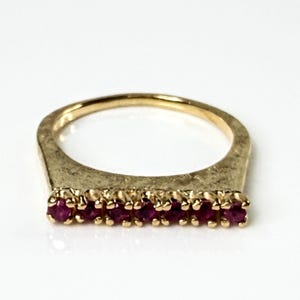 May include: A gold ring featuring a horizontal bar set with eight small, round, deep red rubies. The gold band has a textured, brushed finish across the front bar section, contrasting with the polished, vibrant red gemstones set in a row.