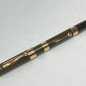 May include: A vintage fountain pen with a dark olive green barrel and a black cap. The pen features gold-colored accents, including a clip and bands. The pen is angled slightly, showcasing its design and craftsmanship.
