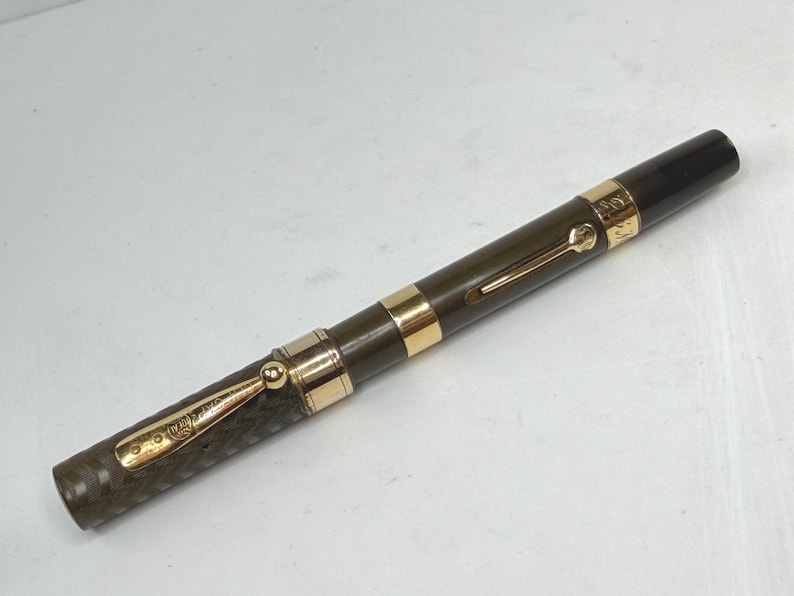 May include: A vintage fountain pen with a dark olive green barrel and a black cap. The pen features gold-colored accents, including a clip and bands. The pen is angled slightly, showcasing its design and craftsmanship.