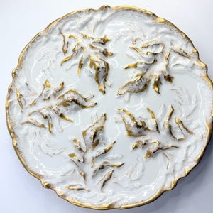 May include: White ceramic plate with a gold rim and embossed leaf design. The plate features a detailed, raised pattern of stylized leaves with gold accents, creating a luxurious and elegant aesthetic. The plate's rim is also trimmed in gold.