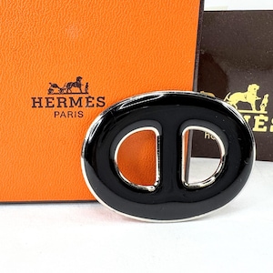 May include: An oval-shaped scarf ring featuring a glossy black enamel finish with a polished silver-tone metal border. The accessory has a central horizontal bar dividing the opening into two distinct sections, creating a sleek and reflective appearance.