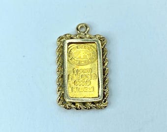 May include: A gold pendant featuring a 1-gram fine gold bar with a 999.9 purity rating. The rectangular bar is set within a gold frame with a rope-like border. The pendant includes the text "1 gram FINE GOLD 999.9 Istanbul".