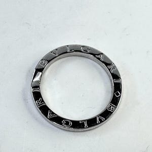 May include: A silver-colored ring with the brand name "BVLGARI" engraved around the outer edge. The ring has a simple, circular design and a polished finish. The letters are evenly spaced around the band.