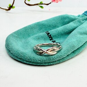 May include: A silver-colored ring with an intertwined knot design rests on a teal drawstring pouch. The pouch has the brand name printed on it. The background is white with a few sprigs of greenery and pink flowers.