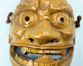 May include: A carved wooden mask with a detailed, expressive face. The mask features a wide open mouth with visible teeth, large eye sockets, and a prominent nose. The wood grain is visible, and it has brown cords attached on each side.