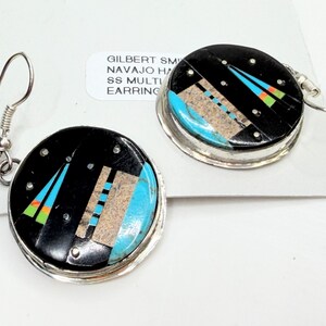 May include: Pair of round earrings with a black background and inlaid turquoise, tan, and colorful geometric designs. The earrings have silver-tone metal trim and ear wires. The text on the tag reads "GILBERT SMITH NAVAJO HANDMADE SS MULTI EARRING."