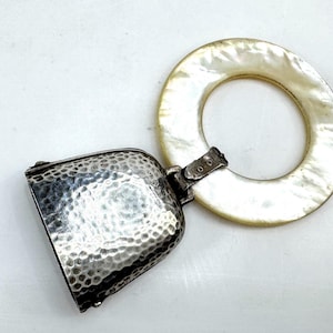 May include: A silver-toned rattle featuring a hammered metal bell attached to a smooth, circular, off-white mother-of-pearl ring. The bell has a textured, dimpled surface and is connected to the ring by a small, hinged metal clasp mechanism.