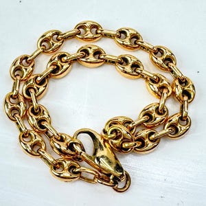 May include: A gold-toned chain bracelet featuring a series of polished, oval-shaped links with a central horizontal bar, connected in a mariner-style pattern. The bracelet is secured with a large, functional lobster claw clasp mechanism.