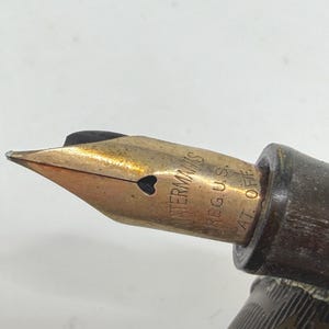 May include: Close-up of a vintage Waterman's fountain pen nib. The nib is gold-colored with a heart-shaped breather hole. The pen's body is dark brown. The text "WATERMAN'S REG. U.S. PAT. OFF." is visible on the nib.