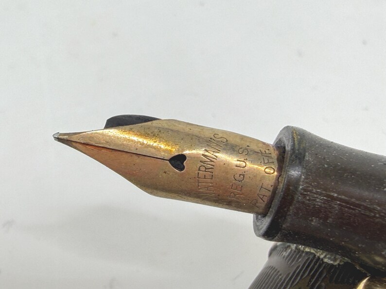 May include: Close-up of a vintage Waterman's fountain pen nib. The nib is gold-colored with a heart-shaped breather hole. The pen's body is dark brown. The text "WATERMAN'S REG. U.S. PAT. OFF." is visible on the nib.