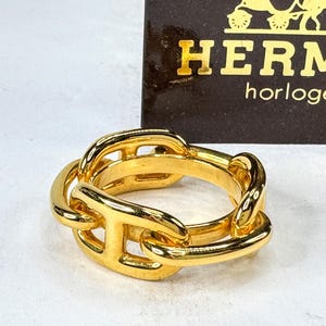 May include: A gold-toned ring featuring a chain-link design with H-shaped links. Behind the ring is a dark brown rectangular card with a gold graphic of a horse-drawn carriage and the text HERMÈS horloger printed in gold lettering.