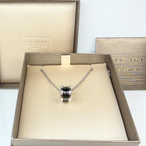 May include: A silver necklace with a cylindrical pendant featuring black and silver accents, presented in a branded box. The box is beige with the brand name embossed on the lid. A matching card is also visible.
