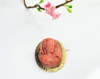 May include: Oval-shaped cameo brooch featuring a carved coral portrait of a woman with flowing hair. The coral cameo is set in a gold-toned, textured frame. A pin is attached to the back for securing the brooch.