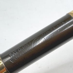May include: Close-up of a vintage fountain pen. The pen has a dark brown barrel with gold-colored accents at each end. The barrel is inscribed with the text "PAT.D.1884 WATERMAN'S FOUNTAIN PEN" and "MAY 23 1899".