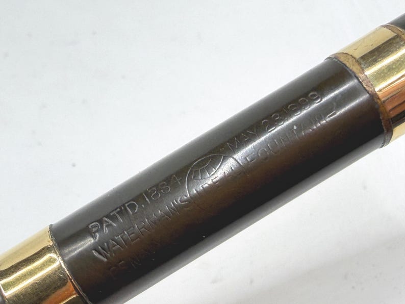 May include: Close-up of a vintage fountain pen. The pen has a dark brown barrel with gold-colored accents at each end. The barrel is inscribed with the text "PAT.D.1884 WATERMAN'S FOUNTAIN PEN" and "MAY 23 1899".