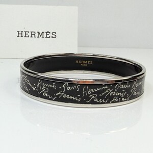 May include: A black and silver Hermes bangle bracelet. The bracelet features white script with the words "Hermes Paris" and a silver trim. A white card with the Hermes logo is in the background.
