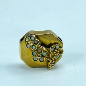 May include: A gold-toned, geometric-shaped jewelry piece with floral accents. The flowers are adorned with small, iridescent opals. The piece has a vintage aesthetic and is set against a light background.