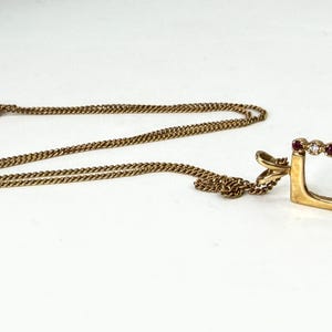 May include: A gold necklace featuring a rectangular pendant frame. The top edge of the pendant is set with alternating small round red rubies and clear white diamonds. The pendant hangs from a delicate gold chain with a standard clasp closure.