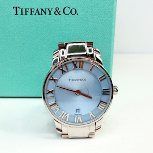 May include: A silver-toned Tiffany & Co. watch with a light blue face and Roman numeral hour markers. The watch has a date display at the 6 o'clock position and a brown second hand. The watch is set against a Tiffany & Co. box.