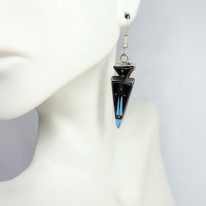 May include: A single black and silver earring with a geometric design is shown. The earring has a triangular shape with a black base, silver accents, and turquoise blue details. The earring is suspended from a silver hook.