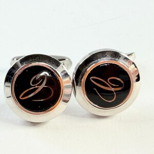 May include: Pair of silver cufflinks with a black center featuring a cursive letter "J" in a copper color. The cufflinks have a polished, reflective finish and a classic, round design.