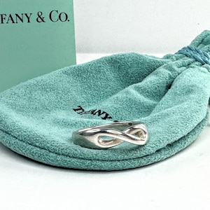 May include: A silver-toned infinity ring rests on a teal drawstring pouch, with a matching box in the background. The ring's design features a continuous loop, symbolizing eternity. The Tiffany & Co. box and pouch add a touch of luxury.
