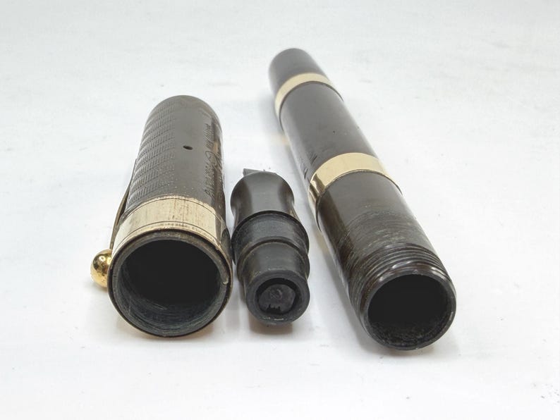 May include: A disassembled fountain pen with a dark brown barrel and cap, accented with gold-colored bands. The pen's nib section is black. The cap has a gold-colored clip and a textured pattern. The pen is lying on a white surface.