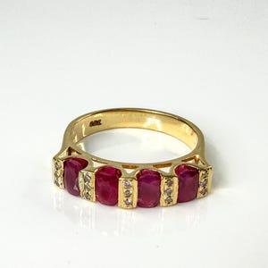 May include: A gold ring featuring four oval-cut red rubies arranged in a row, separated by small vertical bars set with tiny clear stones. The band is polished yellow gold with a visible hallmark stamped on the interior surface of the ring.
