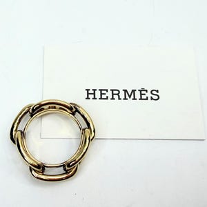 May include: A gold-toned ring with a chain link design. The ring is circular with multiple interlocking links. A white card with the word "HERMÈS" in black lettering is next to the ring.