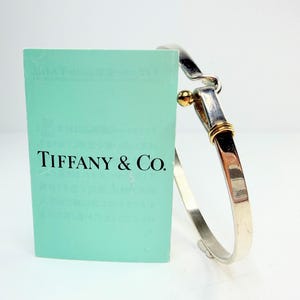 May include: A silver bracelet with gold accents and a clasp, next to a teal card with the Tiffany & Co. logo. The bracelet has a polished finish and a simple, elegant design. The gold accents are visible on the clasp and at the bracelet's joint.