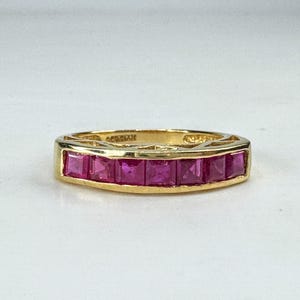 May include: A gold band ring featuring a channel setting with seven square-cut, vibrant pinkish-red gemstones arranged in a horizontal row across the front. The metal band has a polished finish and a visible hallmark stamped on the inner surface.