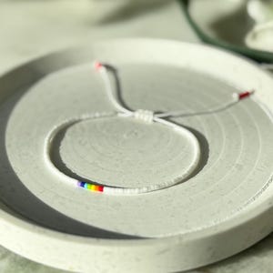 May include: A white beaded bracelet with a rainbow stripe design. The bracelet is on a light gray, circular tray. The bracelet has a sliding knot closure. The background is blurred, with white flowers and green leaves.