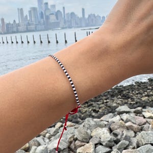以下が含まれることがあります： 赤色の紐留めが付いた白と黒のビーズブレスレットが腕に着用されています。ブレスレットは小さくて丸いビーズでできています。背景には水面、岩の多い海岸線、曇り空の下の都市景観が写っています。
