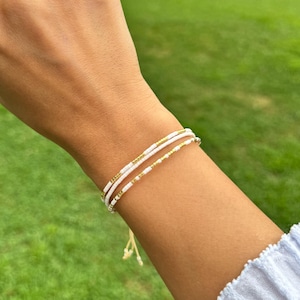 以下が含まれることがあります： 透明な石の付いたゴールドリングとブレスレットをつけた手。ブレスレットは、ゴールドのアクセントが付いた白いビーズが特徴です。背景はぼやけた緑の芝生です。